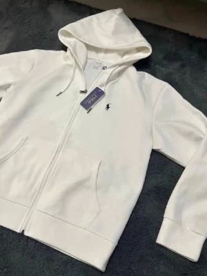 Vit hoodie från Polo Ralph Lauren - Snygg vit hoodie från Polo Ralph Lauren med dragkedja framtill, två fickor och klassisk huva med dragsko. Ikoniska broderade loggan på bröstet. Perfekt för en clean och stilren look. Mjuk bomullsblandning för extra komfort.