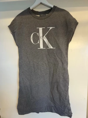 Grå t-shirtklänning från Calvin Klein - Säljer en grå sweat shirtklänning från Calvin Klein med stort vitt CK-tryck på bröstet. Klänningen har korta ärmar och rund halsringning. Perfekt för en avslappnad och stilren look.