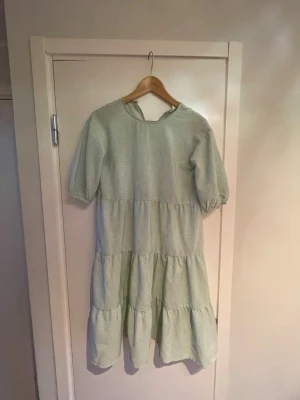 Ljusgrön prickig klänning med volanger - Superfin ljusgrön klänning med små blommor  och volanger i flera lager. Klänningen har halvlånga ärmar och en lös, flowy passform som ger en avslappnad vibe. Perfekt för vår och sommar när du vill känna dig både söt och bekväm.