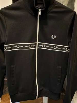 Svart Fred Perry zip hoodie - Svart hoodie från Fred Perry med vit dragkedja och logoband över bröstet. Klassisk design med ståkrage, två fickor fram och broderad Fred Perry-logga på vänster sida. Materialet är mjukt och stretchigt, perfekt för en sportig och stilren look. Skriv för frågor eller fler bilder .