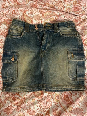 Jeanskjol med fickor från FB Sister - Cool kort jeanskjol från FB Sister i klassisk blå denim. Kjolen har flera stora fickor med knappar och detaljer både fram och bak, samt bälteshällor och dubbla knappar i midjan. Perfekt för en avslappnad och trendig look.