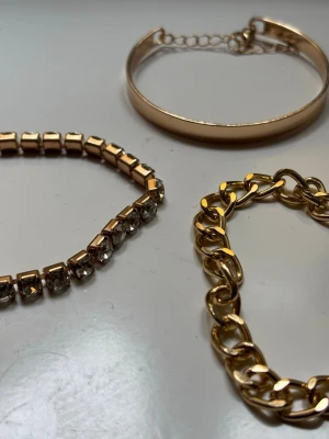 Tre snygga armband i guldton - Säljer tre olika armband: ett chunky kedjearmband i guldfärgad metall, ett stelt armband med kedja för justering och ett armband med fyrkantiga guldfärgade pärlor och glittrande stenar. Perfekta för att mixa och matcha till din stil.