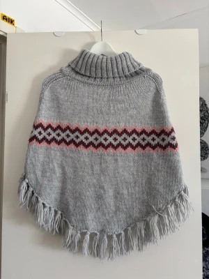 Grå stickad poncho med mönster - Säljer en grå stickad poncho med polokrage och fransar längs kanten🩶storlek 158/170 så typ XS