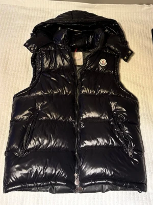 Svart dunväst från Moncler - Snygg svart dunväst från Moncler med glansig finish och klassisk logotyp på bröstet. Västen har dragkedja framtill, två fickor med dragkedja och hög krage. Perfekt för lager-på-lager och streetstyle. Materialet är glansigt nylon med dunfyllning.