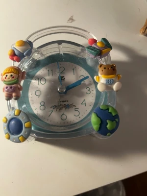 Söt blå väckarklocka med figurer - En blå och transparent väckarklocka i plast med gulliga dekorationer som nalle, jordglob, sol, rymdraket och en liten figur. Klockan har tydliga siffror och visare samt en lekfull design som passar perfekt på skrivbordet eller nattduksbordet.