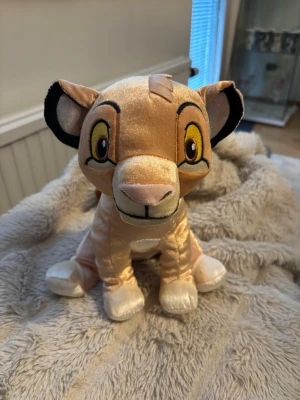 Simba gosedjur från Lejonkungen - Mjukisdjur som föreställer Simba från Lejonkungen. Gosedjuret är ljusbrunt med broderade detaljer, stora gula ögon, svartkantade öron och en liten brun svans. Tillverkad i mjuk plysch och har en klassisk sittande form.