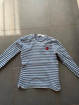 Comme des Garçons longsleeve  - Säljer en långärmad tröja från Comme des Garçons med blå och vita horisontella ränder. Tröjan har en rund halsringning och ett rött hjärta med ögon broderat på bröstet. Perfekt för dig som gillar streetwear och klassisk design. Skick: 9/10, använd ett väldigt fåtal gånger