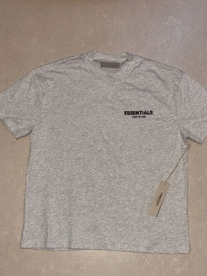Grå Essentials T-shirt Fear of God - Snygg gråmelerad T-shirt från Essentials Fear of God med svart logga på bröstet och stor text på ryggen. Klassisk passform och rund halsringning, perfekt för en clean och stilren look.
