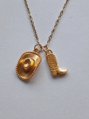 Guldfärgat halsband med cowboytema - Unikt halsband i guldfärg med två berlocker: en cowboyhatt och en cowboyboot. Kedjan är tunn och elegant, och berlockerna har fina detaljer som ger en cool westernkänsla. Perfekt accessoar för dig som gillar statement-smycken och vill sticka ut. Materialet rostfritt stål. 