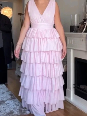 Ljusrosa volangklänning med v-ringning - Säljer en lång, ljusrosa klänning med djup v-ringning och flera lager volanger. Klänningen är ärmlös och har en elegant, romantisk look. Perfekt för speciella tillfällen och ger en riktigt drömmig känsla.   ORGINAL PRIS 2000kr aldrig använd!!