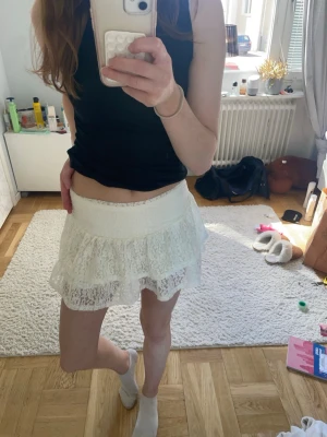 Vit spetskjol med volang - Supersöt vit kortkjol i spets med volangdetaljer. Kjolen har en diskret dragkedja och är perfekt för dig som gillar en romantisk och feminin stil. Passar till både sneakers och sandaler. 
