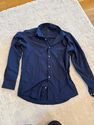 Mörkblå skjorta från Polo Ralph Lauren - Klassisk mörkblå skjorta från Polo Ralph Lauren med vit knappstängning och röd broderad logga på bröstet. Skjortan har lång ärm och normal passform, perfekt för en stilren look.
