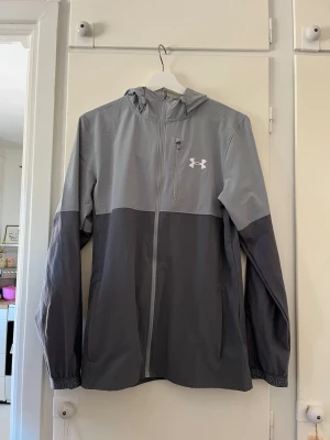 Grå vindjacka från Under Armour - Snygg tvåtonad grå vindjacka från Under Armour med huva och hel dragkedja. Jackan har logga på bröstet, långa ärmar och elastiska muddar med tryckt text. Perfekt för blåsiga dagar och sportiga outfits.