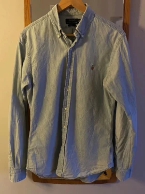 Ljusblå skjorta från Polo Ralph Lauren - Klassisk ljusblå skjorta från Polo Ralph Lauren med button-down krage och broderad logga på bröstet. Skjortan har lång ärm, knäppning framtill och är tillverkad i bomull. Perfekt för en clean och stilren look med den ljusa färgen nu till sommaren. 