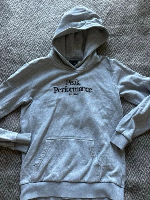 Grå hoodie från Peak Performance - Snygg grå hoodie från Peak Performance med svart broderad logga på bröstet. Klassisk känguruficka framtill, ribbade muddar och mjukt material. Perfekt för en chill och sportig stil. Huvan ger extra mysfaktor och streetkänsla.