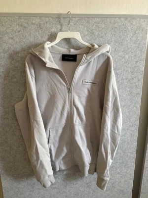 Ljusgrå zip hoodie från Peak Performance - Ljusgrå hoodie med dragkedja från Peak Performance. Klassisk modell med huva, två fickor framtill och ribbade muddar. Diskret logga på bröstet. Perfekt för en avslappnad och sportig stil. Tillverkad i mjukt bomullsmaterial. Om priset inte passar skicka förslag 
