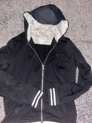 Svart hoodie med dragkedja från Guess - Svart hoodie från Guess med dragkedja framtill och grå insida på huvan. Tröjan har ribbade muddar med vita ränder och klassisk passform. Perfekt för en chill och stilren look. Materialet är mjukt och bekvämt, troligen bomullsblandning. 
