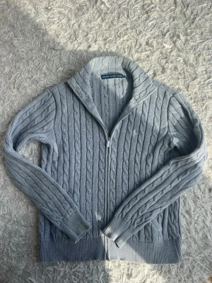 Grå kabelstickad ziptröja Polo Ralph Lauren - Kabelstickad grå tröja med dragkedja från Polo Ralph Lauren. Tröjan har hög krage, långa ärmar och ribbade muddar samt nederkant. Ska vara ärlig den har krympt, påverkar inte materialet eller proportionerna, men sitter mer som en S-M nu. Kan skicka mått som så önskas