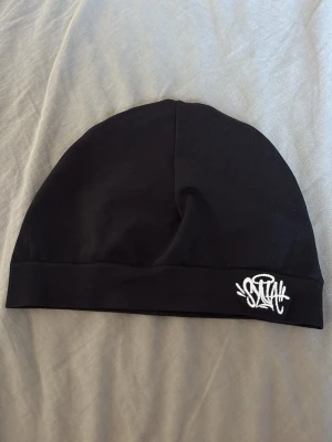 Svart mössa med broderad logga - Svart, stilren mössa i mjukt och stretchigt tyg med broderad vit Syna-logga framtill. Enkel design som passar perfekt till streetwear och vardagsoutfits. Mössan har en klassisk passform och är lätt att matcha med olika stilar.