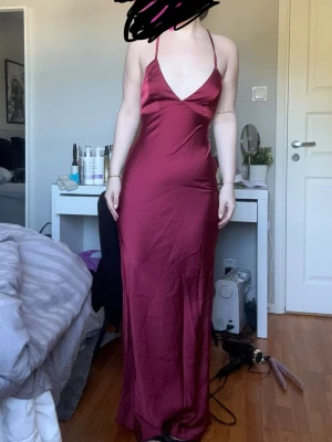 Vinröd långklänning med öppen rygg - Säljer en vinröd långklänning i satin material med smala axelband och djup v-ringning. Klänningen har öppen rygg med snörning och en elegant, figurnära passform.❤️Aldrig använd men sätter ”Mycket bra skick” för den har ingen prislapp!