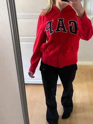 Röd hoodie med dragkedja från GAP - Snygg röd hoodie från GAP med dragkedja och stor logga framtill. Motsvarar storlek S. 