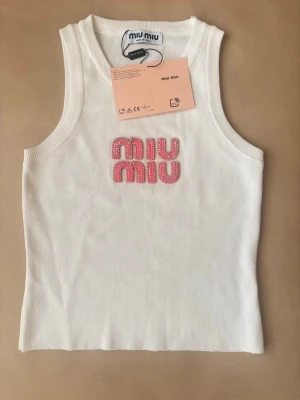 Vit linnetopp från Miu Miu - Snygg vit linnetopp från Miu Miu med rosa pärlbroderad logotyp på bröstet. Toppen är ärmlös och har en ribbad struktur som ger en modern och clean look. Perfekt för att skapa en trendig outfit.