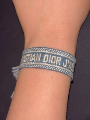 Blått Christian Dior J'Adior armband - Säljer ett snyggt blått tygarmband från Christian Dior med texten 'J'Adior' broderat i vitt. Armbandet har en bohemisk känsla med tofsar i ändarna och är justerbart för att passa de flesta handleder. Perfekt accessoar för att lyfta din outfit.