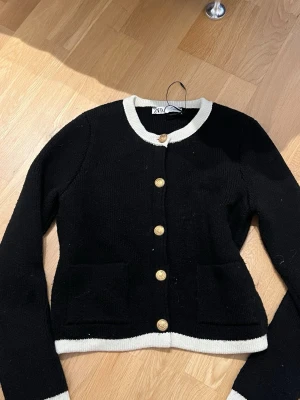 Svart kofta med vita kanter från ZARA - Stilren svart kofta från ZARA med vita kontrastkanter vid hals, ärmslut och nederkant. Koftan har rund halsringning, guldknappar framtill och två fickor. Använd fåtal gånger 