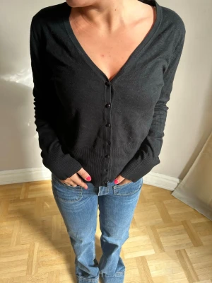 Svart cardigan med v-ringning💖💖 - Svart cardigan med djup v-ringning och knäppning framtill. Långa ärmar och ribbade muddar vid ärmslut och nederkant. Enkel och stilren design som passar till det mesta. Perfekt att slänga över en t-shirt eller linne.