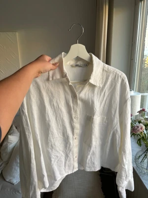 Vit linneskjorta från Zara - Säljer en vit, croppad skjorta från Zara i luftigt linne. Skjortan har klassisk krage, knappar framtill och en bröstficka. Perfekt för en avslappnad och trendig look.