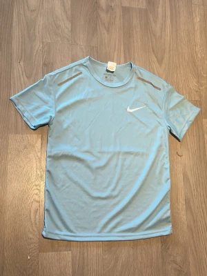 Ljusblå Nike tränings-tshirt - Ljusblå tränings-tshirt från Nike med vitt swoosh-tryck på bröstet. Tillverkad i ett lätt och ventilerande funktionsmaterial, perfekt för träning. T-shirten har rund halsringning och korta ärmar samt diskreta reflexdetaljer på axlarna.