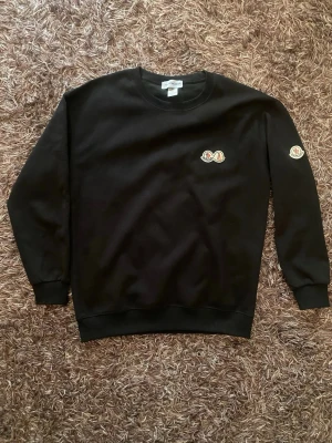 Svart Moncler sweatshirt med logga - Svart sweatshirt från Moncler med rund halsringning och ribbade muddar. Tröjan har två broderade Moncler-loggor, en på bröstet och en på ärmen. Tillverkad i mjukt bomullsmaterial för en clean och stilren look.