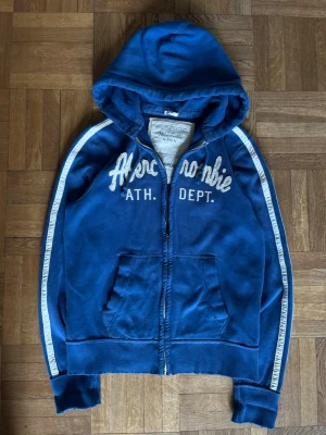 Blå hoodie från Abercrombie & Fitch - Blå zip-hoodie från Abercrombie & Fitch med broderad vit text på bröstet och vita ränder längs ärmarna. Mjuk fleece på insidan, två fickor fram och dragsko i huvan. Perfekt för en avslappnad och sportig stil.