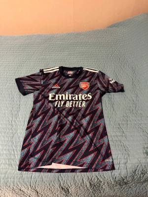 Arsenal Adidas matchtröja zigzag - Snygg Arsenal matchtröja från Adidas med mörkblå bas och rött/blått zigzag-mönster. Tröjan har korta ärmar, rund hals och vita Adidas-ränder på axlarna. Arsenal-logga och Emirates Fly Better-tryck på bröstet. Perfekt för fotboll eller träning.