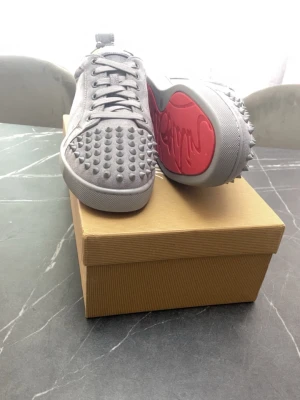 Grå Christian Louboutin sneakers med nitar - Säljer ett par gråa sneakers från Christian Louboutin med ikoniska röda sulor och coola nitar över hela tån och sidorna. Skorna har snörning och är tillverkade i mockaliknande material. Perfekta för dig som vill sticka ut med en edgy och lyxig stil.