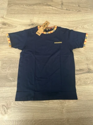 Marinblå Burberry t-shirt med ränder - Snygg marinblå t-shirt från Burberry med klassiska randiga detaljer i beige, svart och rött vid ärmslut och halsringning. T-shirten har korta ärmar och en liten Burberry-logga broderad på bröstet. Perfekt för en trendig och stilren look.