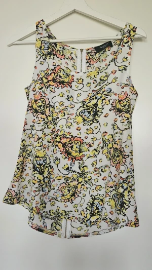 Blommig linnetopp från Sisters Point - Snygg ärmlös topp från Sisters Point med blommigt mönster i gult, rosa och svart på vit botten. Toppen har rund halsringning och dragkedja  bak för en cool detalj. Perfekt för dig som gillar färg och mönster. Använd endast ett fåtal gånger.