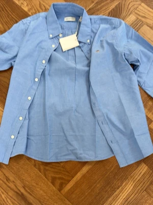 Blå skjorta från Gant med knappar - Klassisk blå skjorta från Gant med button-down krage och vita knappar. Skjortan har lång ärm och liten broderad logga på bröstet. Tillverkad i bomull och passar perfekt till en stilren look.