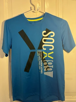 Blå tränings-tshirt från SOC - Säljer en blå tränings-tshirt från SOC med coolt tryck i svart, vitt och gult på framsidan. T-shirten har rund hals och är tillverkad i ett lätt och funktionellt material som passar perfekt för träning och sportaktiviteter.