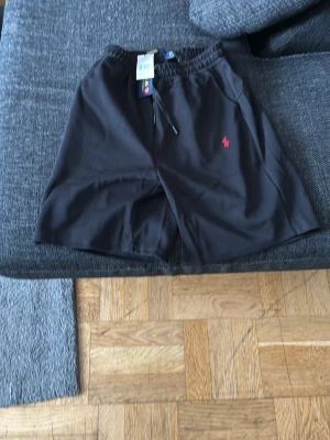 Svarta shorts från Polo Ralph Lauren - Svarta shorts från Polo Ralph Lauren med elastisk midja och snörning. Klassisk röd logga broderad på vänster sida. Tillverkade i mjukt material som känns skönt mot huden. Perfekta för en avslappnad och sportig look.