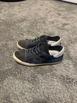 Mörkblå och grå sneakers från Philippe Model - Snygga sneakers i mocka och textil från Philippe Model. Skorna är mörkblå och grå med vita detaljer vid hälen och har en klassisk låg profil. Perfekta för dig som gillar stilrena och bekväma sneakers med en sportig touch.