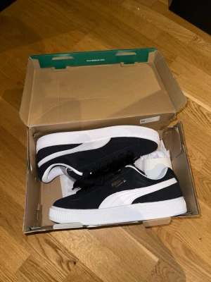 Svarta Puma Suede sneakers - Aldrig använda och stilrena svarta Puma Suede sneakers med vit sula och vit sidodetalj. Skorna har breda svarta skosnören, finns vita snören i lådan också. Ovandelen är i mocka och insidan är vit med gröna detaljer på plösen. Perfekta för en trendig streetstyle-look.