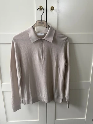 Beige ziptröja från Tiger of Sweden - Snygg beige långärmad tröja från Tiger of Sweden med krage och halv dragkedja framtill. Tillverkad i mjukt material som känns lyxigt och stilrent. Perfekt för dig som gillar minimalistisk och clean stil.