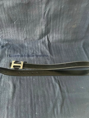 Svart skärp med H-spänne  H’e’r’m’è’s - Stilrent svart skärp från H’e’r’m’è’s med klassiskt H-formad spänne i silverfärg. Skärpet har vital kontrastsömmar längs kanterna. Perfekt accessoar för att lyfta din outfit.