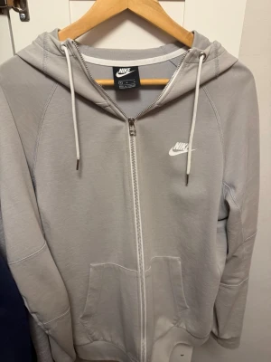 Ljusgrå zip-hoodie  Nike - Snygg ljusgrå hoodie från Nike med dragkedja framtill, huva med vita snören och klassisk logga på bröstet. Mjuk bomullsblandning och två fickor framtill. Perfekt för en avslappnad streetstyle-look.