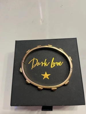 Guldfärgat armband från Dark Love - Coolt guldfärgat armband från Dark Love med nitar runt hela bandet. Armbandet har en rund form och är gjort i metall med en blank finish. Perfekt accessoar för att ge din outfit en edgy touch.