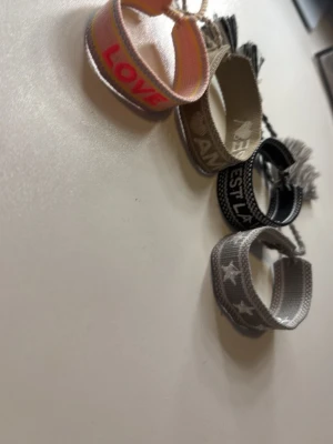 Broderade tygarmband med text - Fyra breda tygarmband i olika färger med broderade texter och mönster. Ett armband är ljusrosa med ordet LOVE i rött, de andra har beige, svart och grå toner med olika texter och stjärnor. 