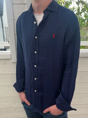 Mörkblå linneskjorta från Ralph Lauren - Snygg mörkblå skjorta från Ralph Lauren i linne med klassisk krage och vita knappar. Skjortan har lång ärm och den ikoniska röda loggan broderad på bröstet. Perfekt för en stilren och avslappnad look.