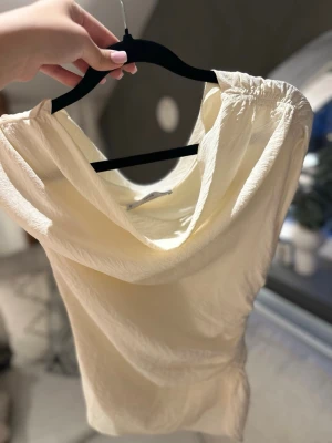 Krämvit asymmetrisk topp från Mango  - Säljer en krämvit asymmetrisk topp från mango med draperad halsringning. Den har en dragkedja på sidan som gör det lättare att sätta på och ta av sig den. Endast använd ett fåtal gånger.