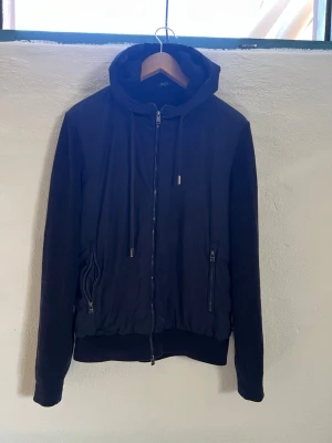 Hugo boss cardigan  - Stilren mörkblå cardigan med huva och dragkedja framtill. Jackan har ribbade ärmar och muddar, samt två fickor med dragkedja. Perfekt för dig som gillar en clean look och vill ha något enkelt men snyggt till vardags. Materialet ser ut att vara en mix av bomull och polyester. Mkt bra skick 